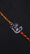 A close-up of a traditional Indian Rakhi bracelet featuring a silver-toned metal nameplate engraved with 'Mohan' and 'Aastha', embellished with small clear stones, threaded on a braided red and yellow cord against a dark background.